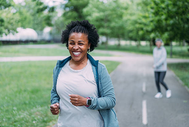 Smiling woman jogging on a park path, with another blurred runner behind her on a green, tree-lined day.