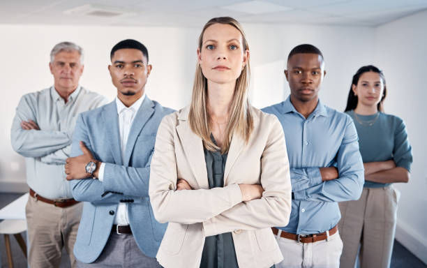 Business team of five standing confidently with arms crossed in a bright office setting, wearing neutral and blue tones, serious mood.