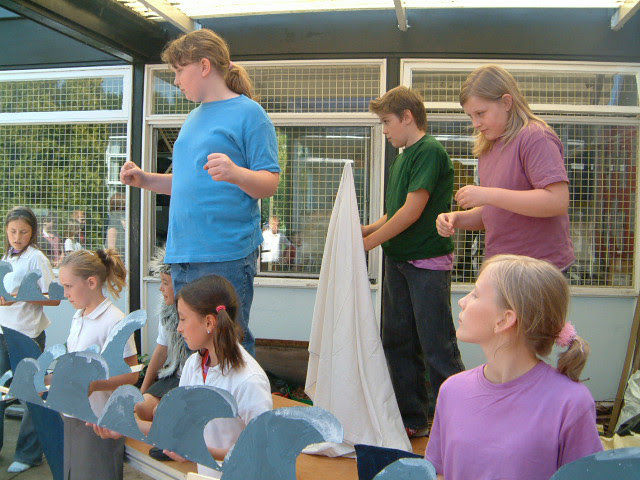 Children perform on a small stage with cardboard cutouts, standing and sitting in colorful shirts under a covered outdoor area.