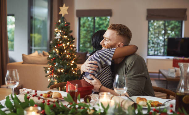A couple hugs at a festive table with a Christmas tree in the background. Warm lighting, holiday decor, and wrapped gifts create a joyful mood.