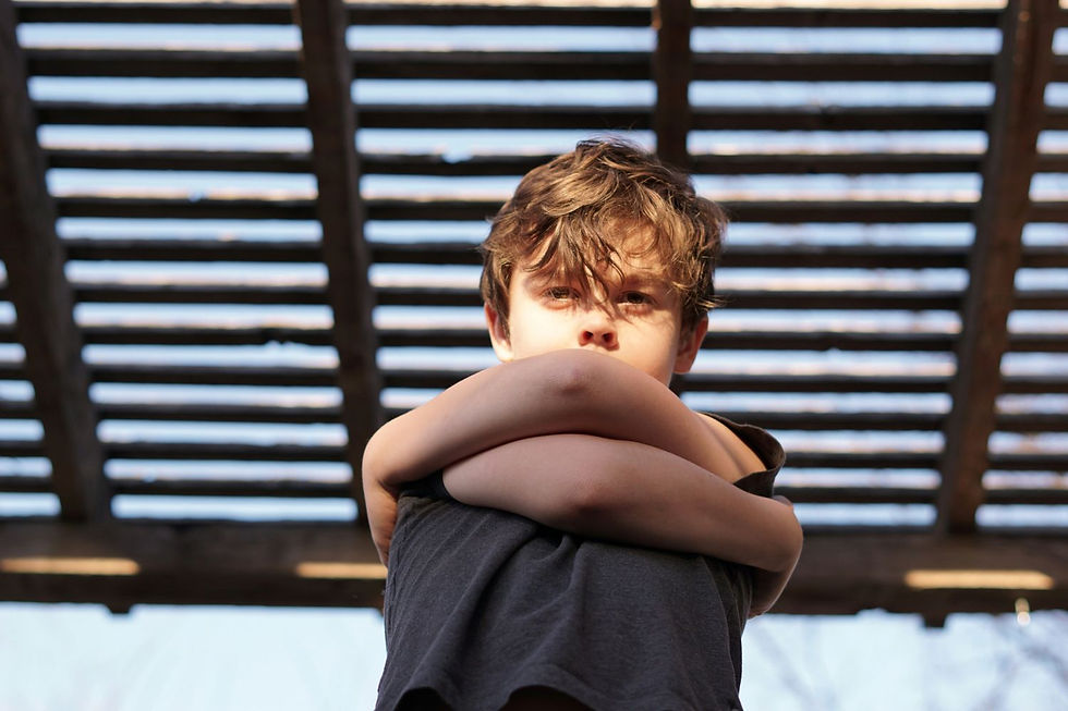 A young boy stands with his arms crossed, looking down with a serious expression under a slatted wooden roof structure.