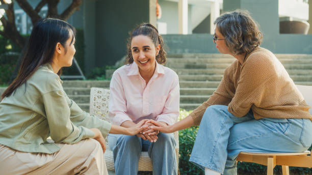 Three women sit outdoors, smiling and holding hands. One wears a pink shirt, another a beige sweater. Greenery and steps in the background.