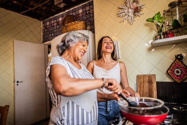 Two women laughing while cooking in a cozy kitchen. One stirs a red pan. Tiled walls, plants, and decorative items are visible. Warm mood.