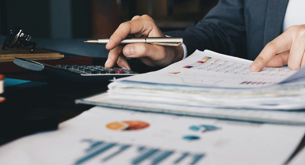 Hands with pen and calculator reviewing financial charts and paperwork at a desk in an office, focused mood.