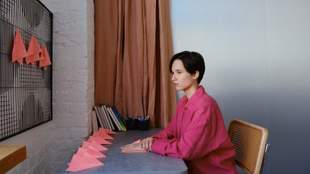 Person in a pink shirt sits at a desk, folding pink paper shapes. Books and pencils are in the background. Neutral, focused mood.