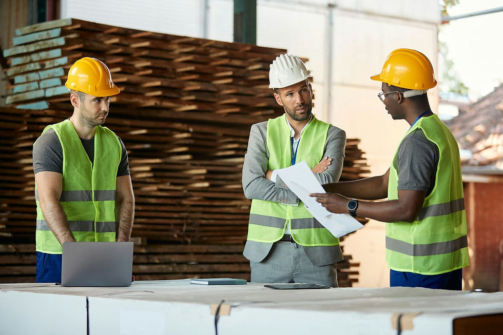 Three construction workers in hard hats and safety vests review plans beside a laptop, with stacked lumber in the background.