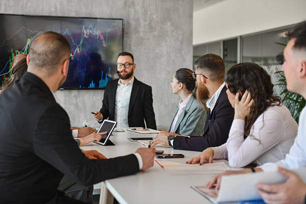 Business meeting with presenter beside a chart screen, team seated at a table in a modern office, focused and attentive