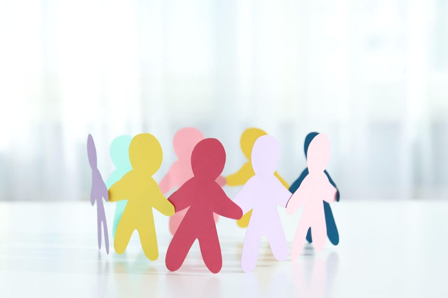 Colorful paper cutout people standing together on a bright white surface, softly lit background, suggesting teamwork and unity