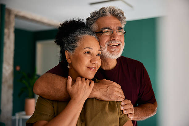 Older couple embracing, smiling in a room with teal walls and a potted plant. The atmosphere is warm and affectionate.