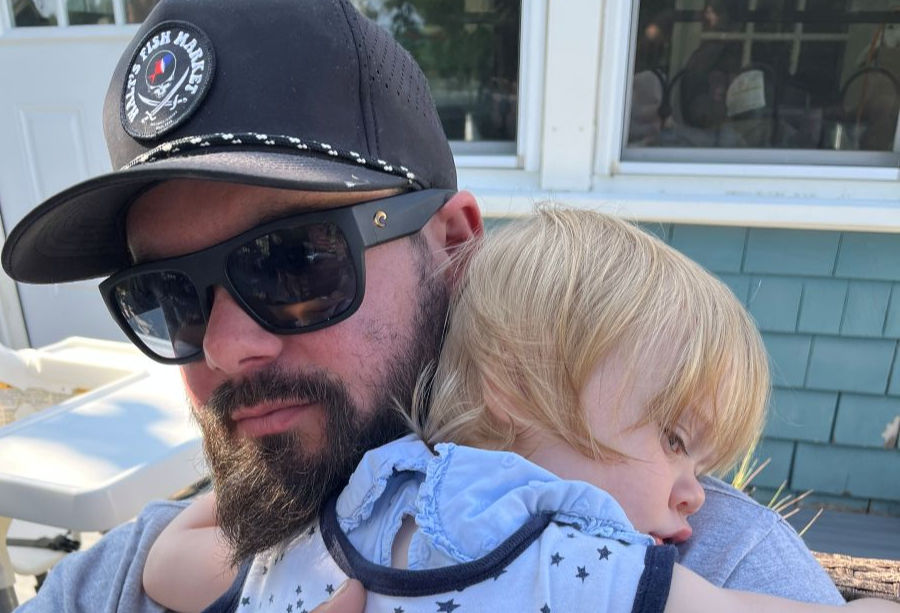 Man in sunglasses and cap holding a sleeping child. They are outdoors near a blue house. The cap has "Phil's Fish Market" text. Peaceful scene.