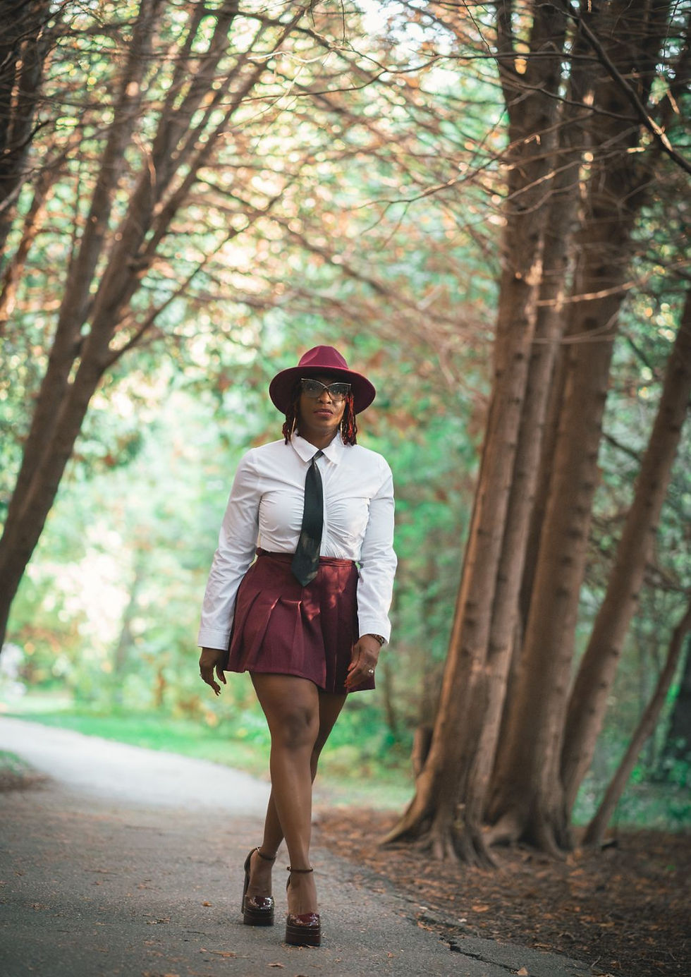 Person in white shirt, tie, and burgundy skirt walks confidently through a forest path. Wears burgundy hat and glasses. Sunny ambiance.
