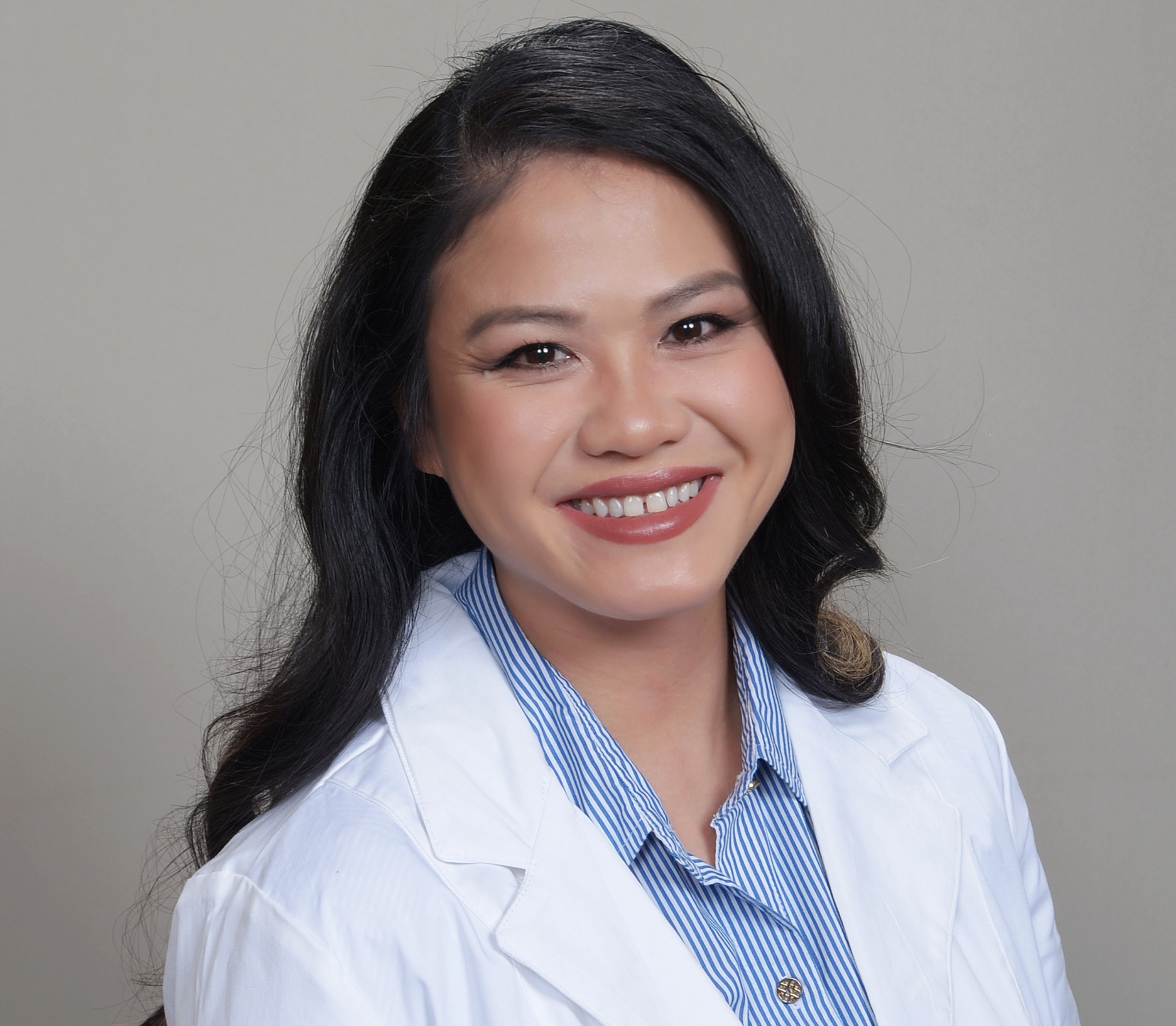 Smiling woman in a white lab coat and blue striped shirt posed against a plain gray background, warm and professional.