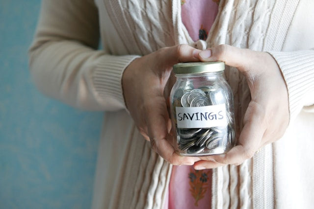A person is holding a jar labeled "SAVINGS" filled with coins.