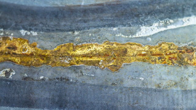 Close-up of textured metal surface with a rusty gold streak across a gray-blue background. The image has an industrial and aged look.