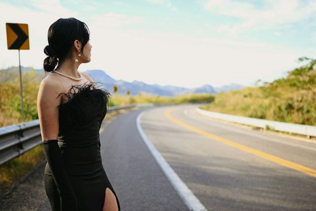 Woman in a black strapless dress stands by a curving mountain road, looking left beside a yellow turn sign under a bright sky