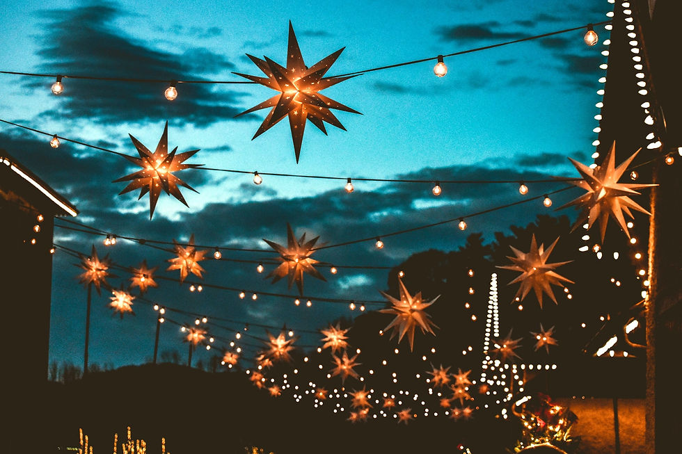 Festive star-shaped lights and bulbs hang across a street at dusk, with a blue sky and clouds in the background creating a warm ambiance.
