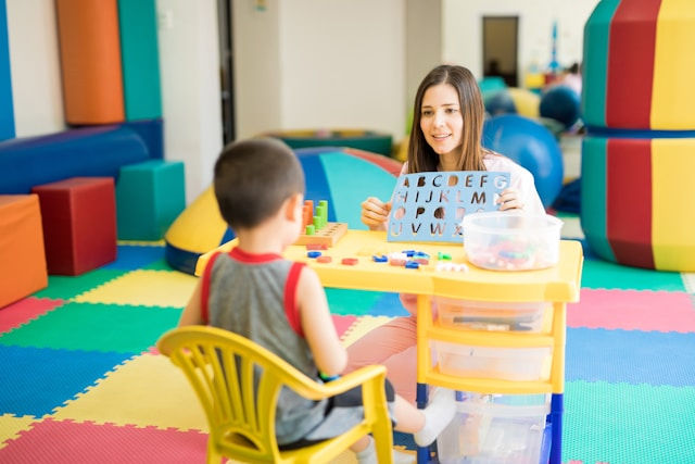 Woman showing alphabet chart to a child in a colorful preschool, with foam mats and toys. Bright, educational setting with a cheerful mood.