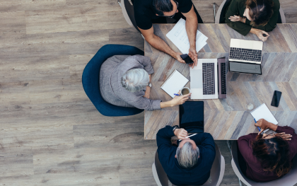 Overhead view of four coworkers around a wooden table with laptops and papers in a focused office meeting.
