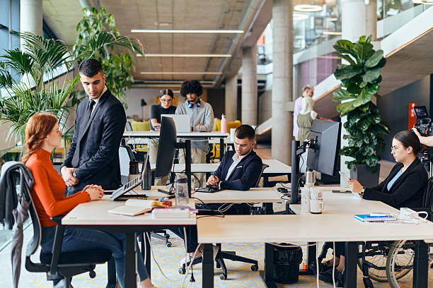 Open office scene with diverse people working at desks. Laptops, plants, and notepads visible. Bright, modern setting with a collaborative mood.