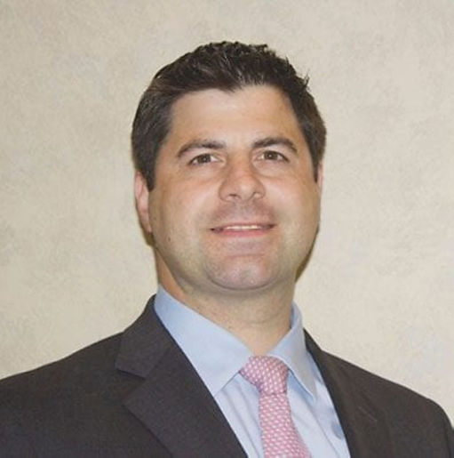 Portrait of a man in a dark suit, light blue shirt, and pink tie, standing against a plain beige wall with a neutral expression.