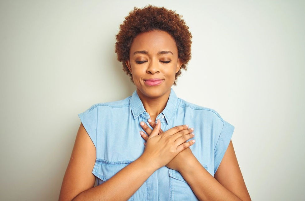 A person with short curly hair, eyes closed, and hands on chest, wears a blue shirt, against a neutral background, expressing calm.