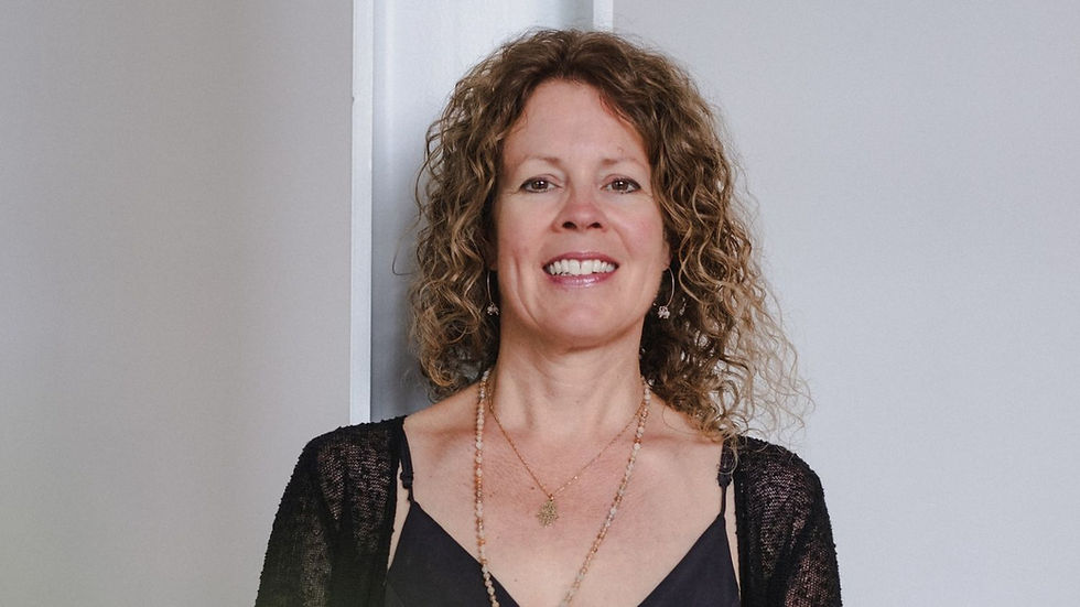 Smiling woman with curly hair, wearing a black top and necklace, against a light grey background. Relaxed and joyful mood.