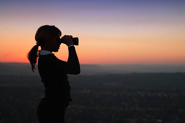 A silhouette of a woman looking through binoculars at sunset, standing against a colourful sky.