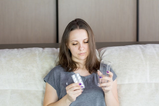 Woman sitting on a couch, holding a glass of water in one hand and a blister pack of pills in the other. Neutral expression, beige background.