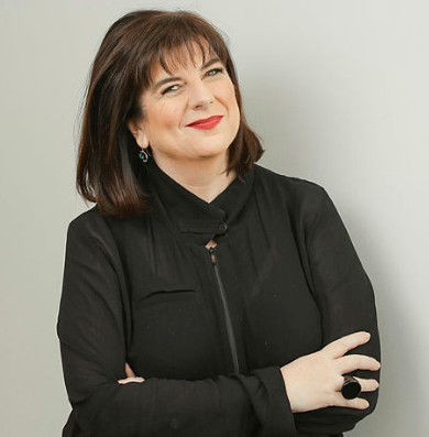 Woman in black top smiling with arms crossed, against a plain light gray background. She has short brown hair and wears a black ring.