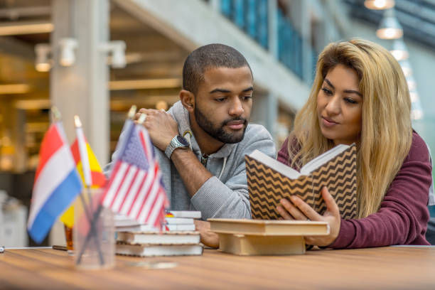 A man and woman read together at a table decorated with international flags, books, and a notebook. Background shows a modern library setting.