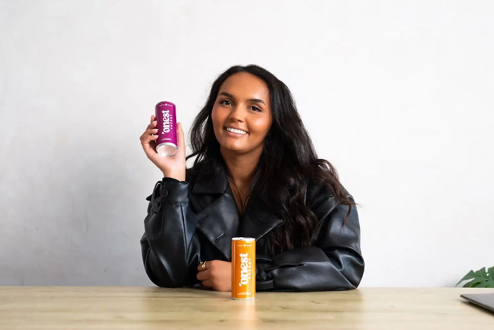 Woman in a black leather jacket smiles, holding a purple can labeled "onest"; an orange can sits on the table. Minimalist background.