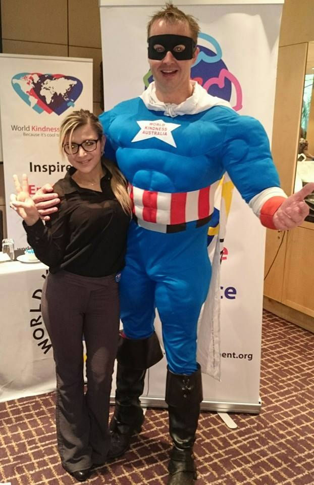 A person poses with a man in a superhero costume labeled "World Kindness Australia." A banner and table with plates are in the background.