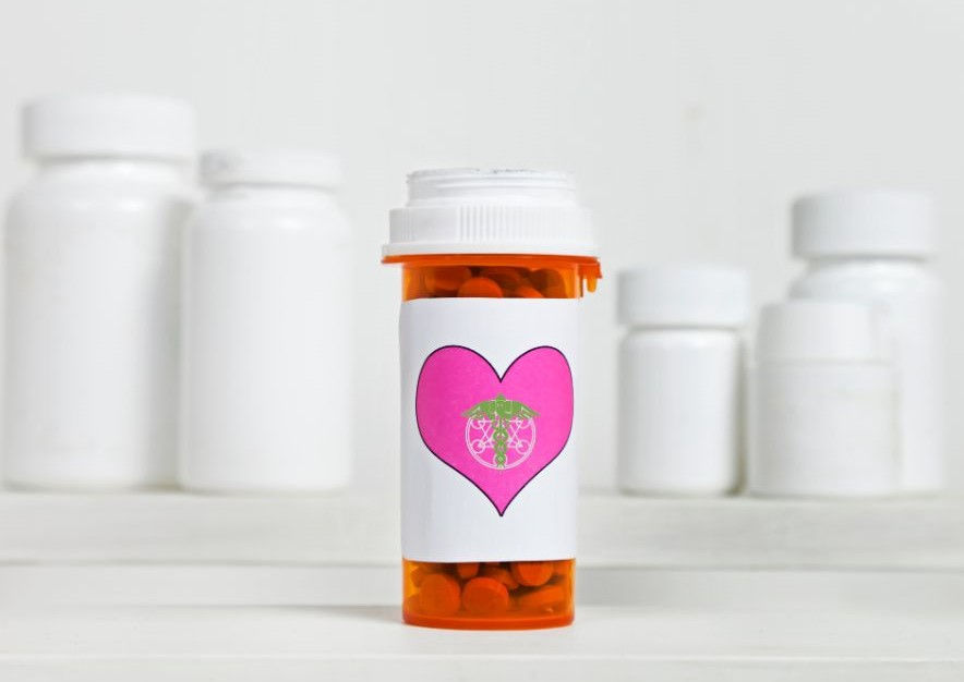 Orange pill bottle with pink heart label, featuring a green medical symbol, in front of blurred white bottles on a white background.