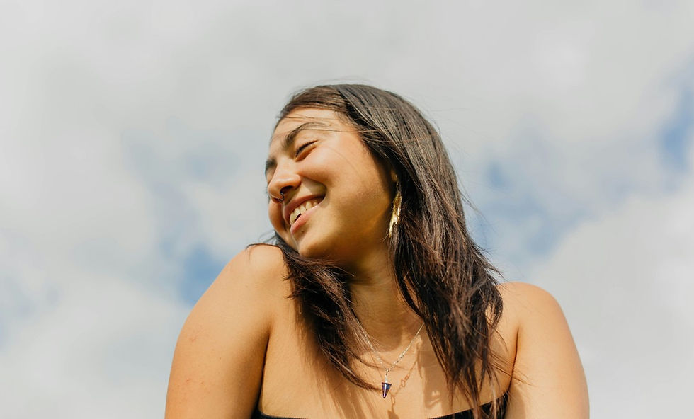 Smiling young woman outdoors against a cloudy sky, head turned aside, wearing a necklace and earring.