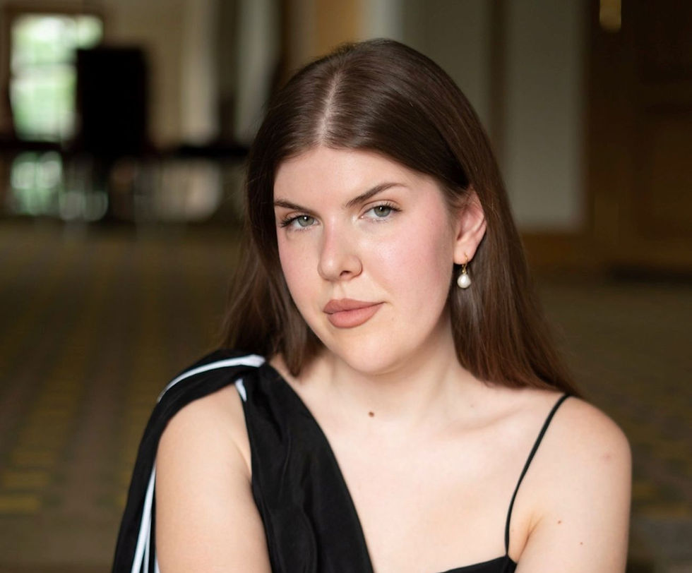 Portrait of a young woman indoors in a black dress and pearl earring, looking calmly at the camera, blurred room behind.