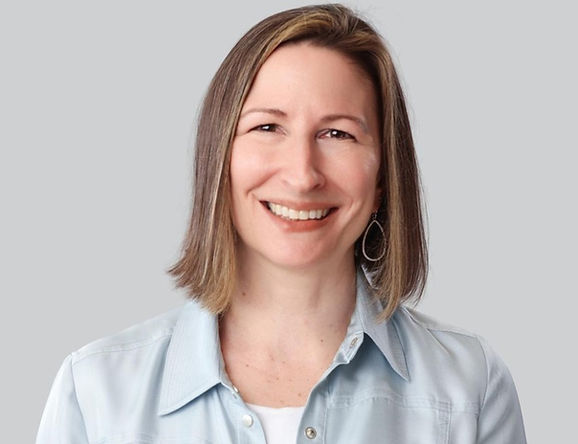 Smiling woman in a light blue shirt against a plain gray background, posed for a portrait.