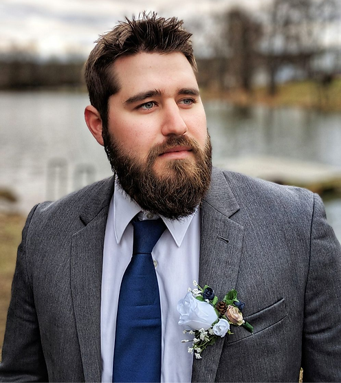 Man in a gray suit and tie with a floral boutonniere on his left lapel, standing near the camera and gazing into the distance beside a calm lakeside backdrop.