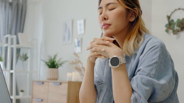 Woman meditating with closed eyes in a serene room. Wearing a blue shirt and smartwatch, surrounded by plants and soft decor.