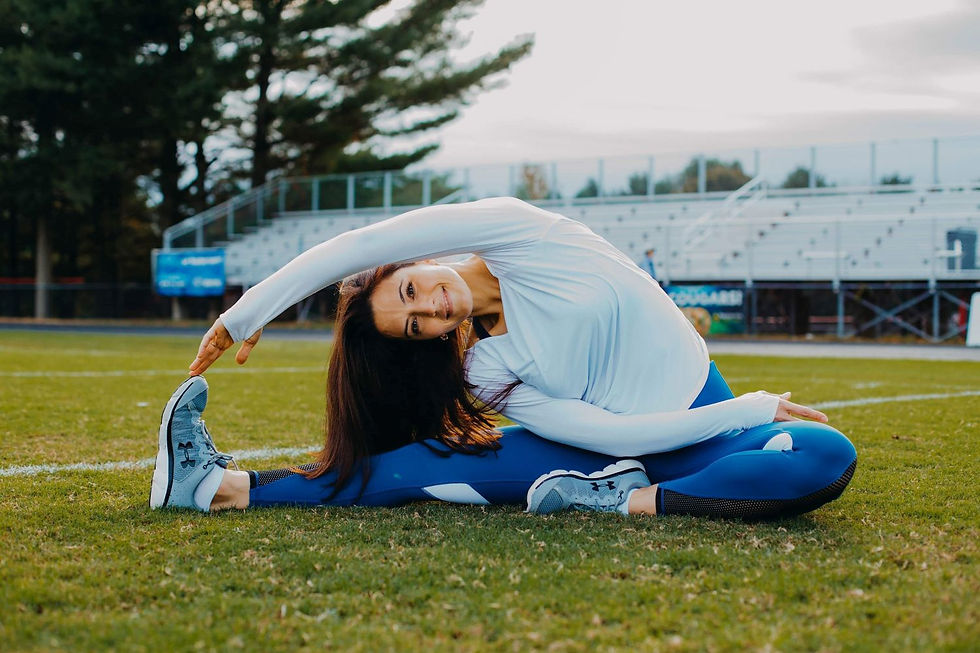 Woman in white top and blue leggings doing a side stretch on a grassy sports field, with bleachers and trees behind her.