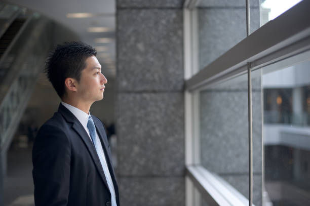 Man in a suit gazes thoughtfully out a large window in a modern office space, with gray walls and ambient lighting.