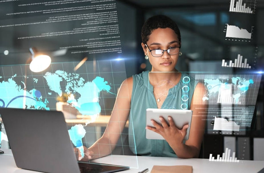Focused woman using tablet and laptop with holographic world maps, charts, and data overlays in a modern office setting.