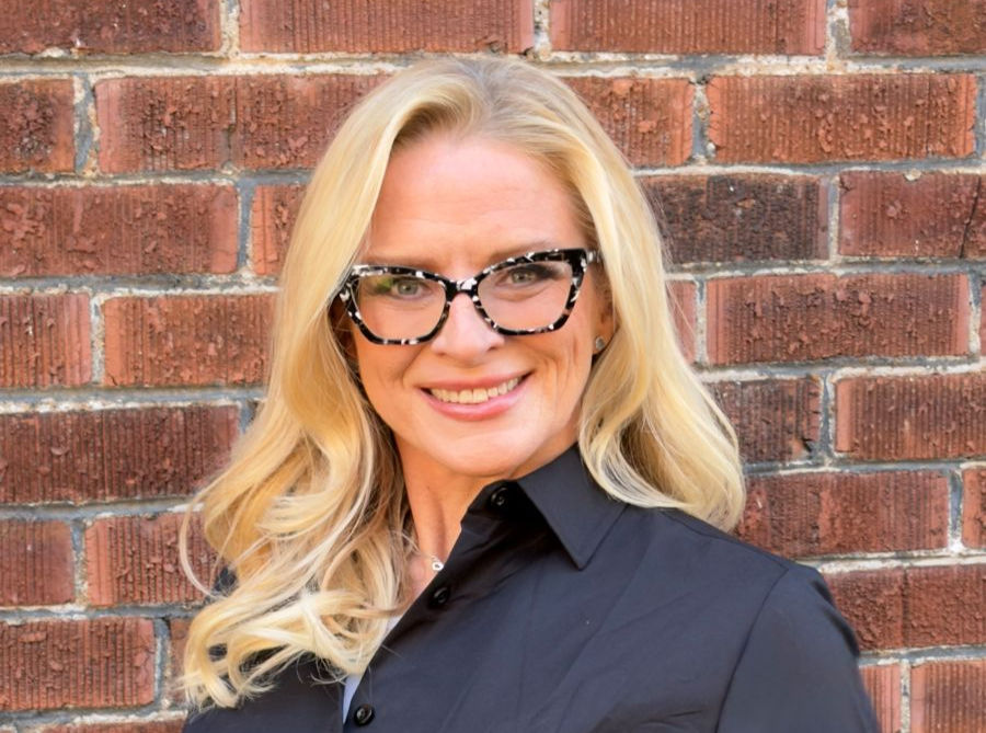 Smiling woman with blonde hair and patterned glasses wearing a black shirt, set against a brick wall background.