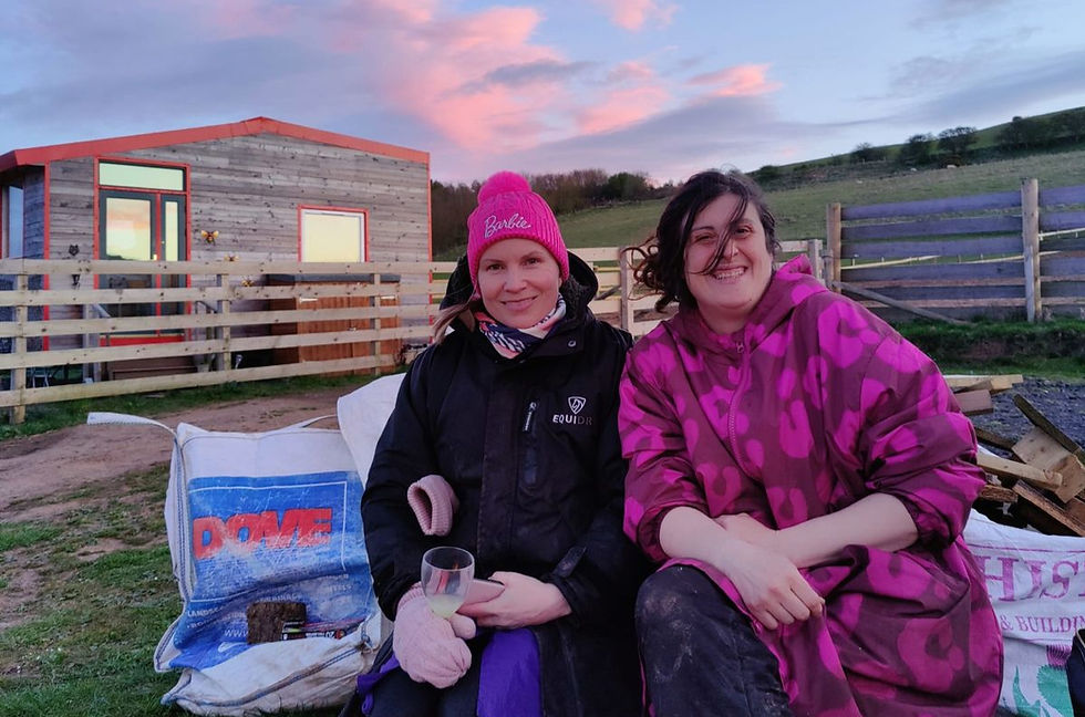 Two smiling women sit on cushions outdoors at dusk; one wears a pink Barbie beanie and the other a magenta coat, with DOME visible.