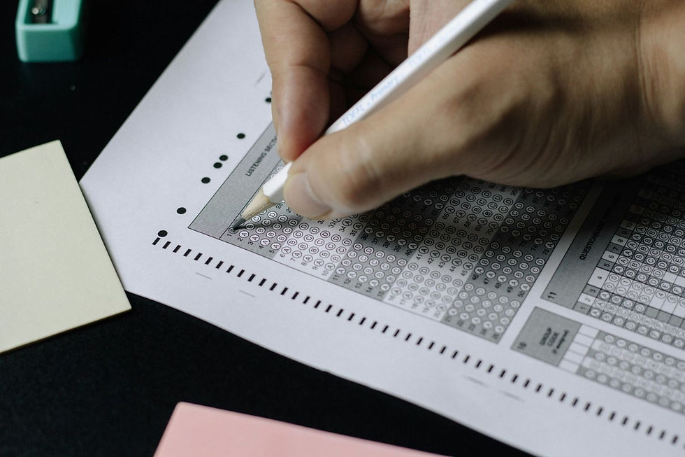 Hand filling multiple-choice exam sheet with a pencil on a dark desk, surrounded by pastel-colored papers.