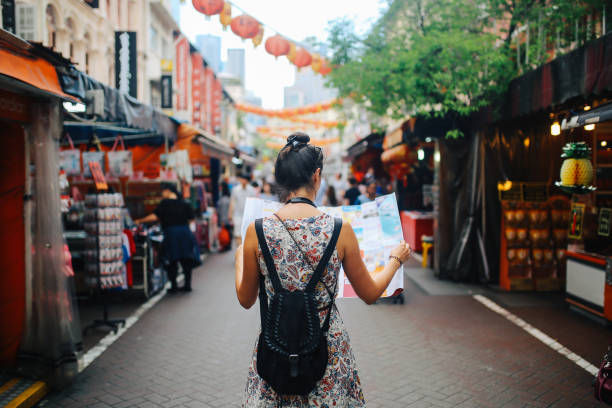 A woman with a backpack holds a map in a vibrant market street with lanterns above. She wears a floral dress, exploring during the day.