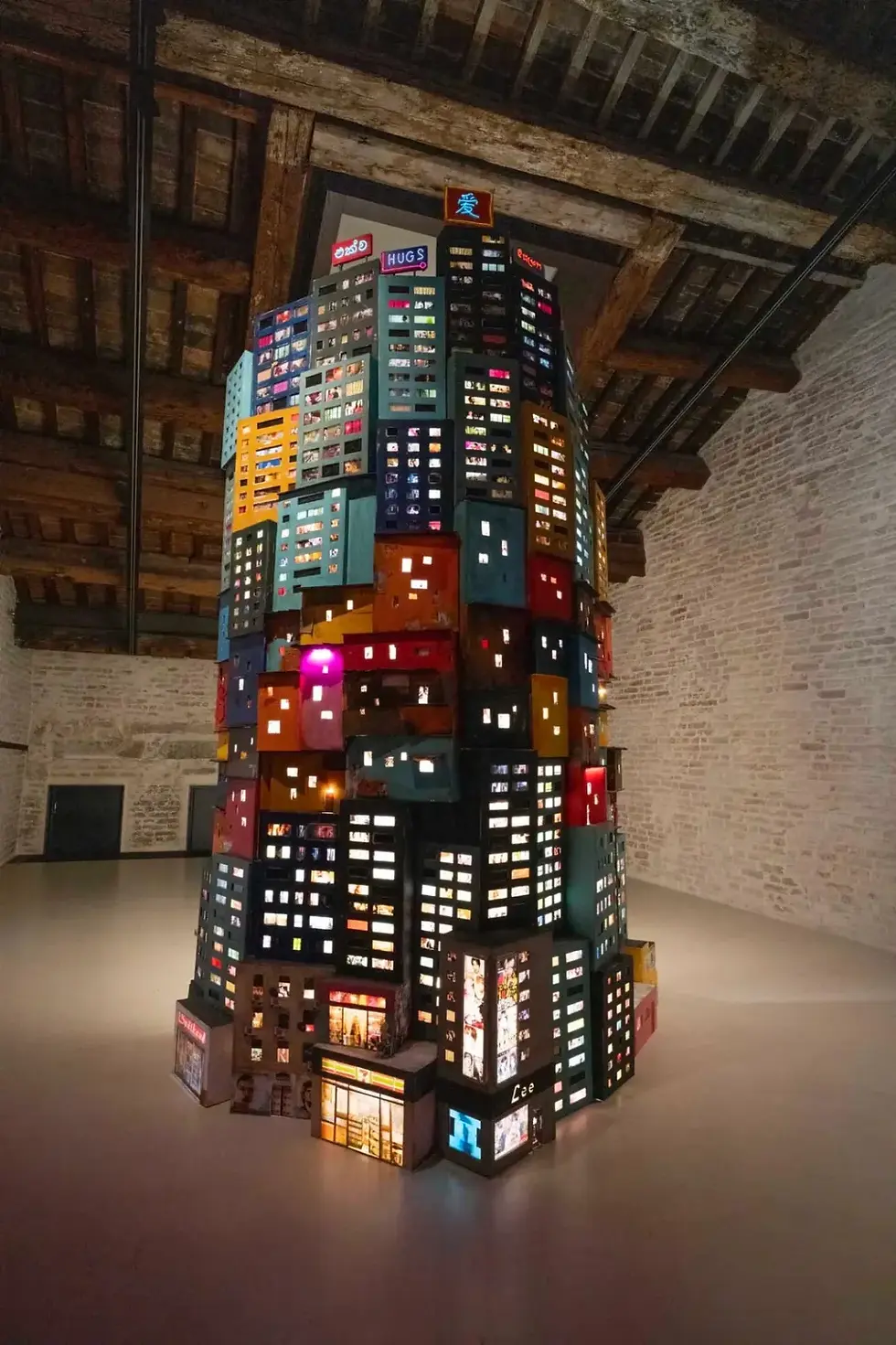 Illuminated tower of colorful miniatures resembling buildings, inside a rustic room with wooden beams and brick walls. Signs: HUGS, Lee.