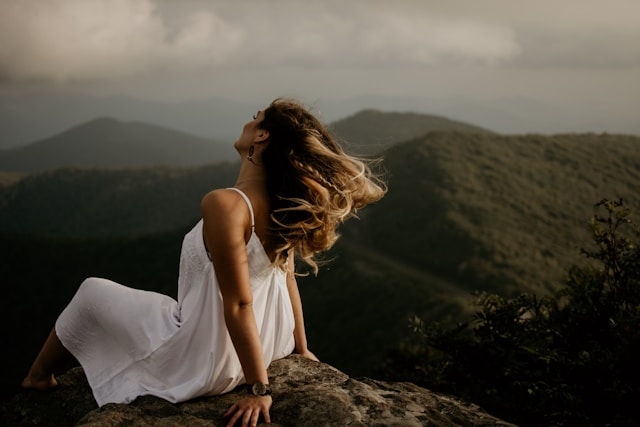 A woman in a flowing white dress sitting on a rock, facing away, with her hair blowing in the wind against a backdrop of rolling green mountains under a cloudy sky.