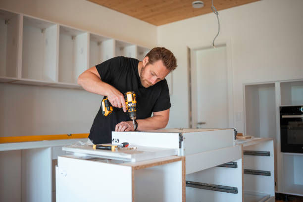 Man in black shirt using a cordless drill to assemble a white kitchen cabinet in a modern unfinished room.