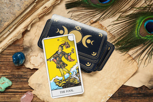 Tarot card "The Fool" on a wooden table with peacock feathers, colored stones, and a decorative card deck. Mystical and vibrant setting.