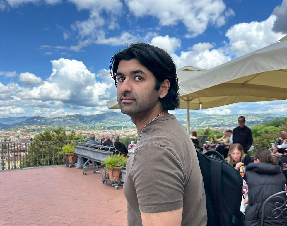 Man with a backpack looks over shoulder on a terrace with scenic views. People seated at tables, cloudy sky above, creating a relaxed mood.